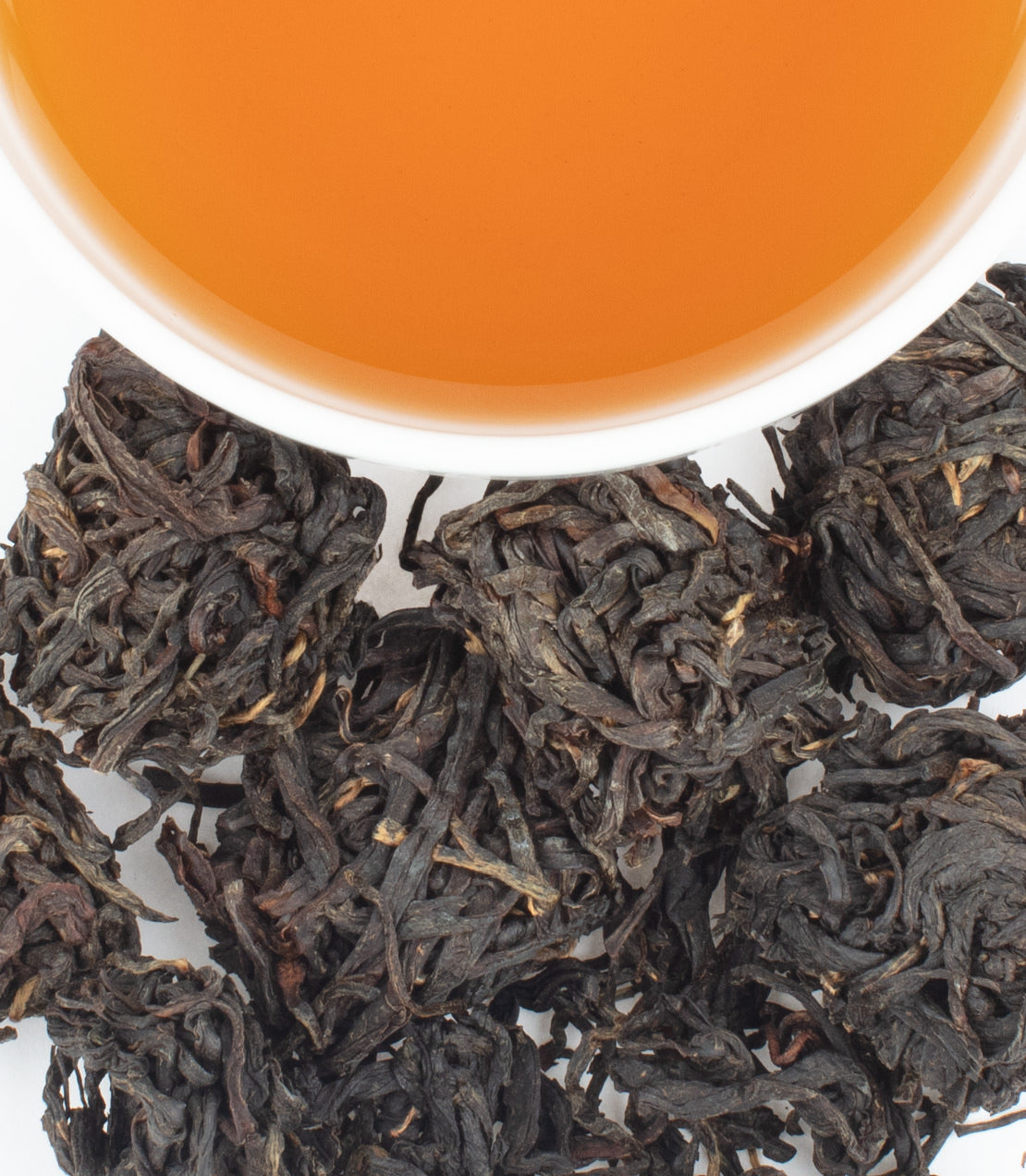 A cup of amber tea sits above a pile of dark, twisted tea leaves, some bunched into cubes
