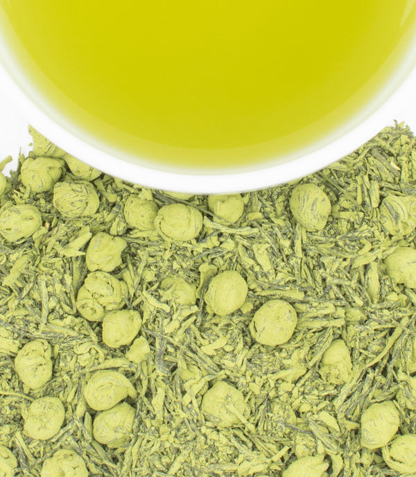 A white cup filled with bright green tea sits above a pile of loose green tea leaves and rolled green tea pearls