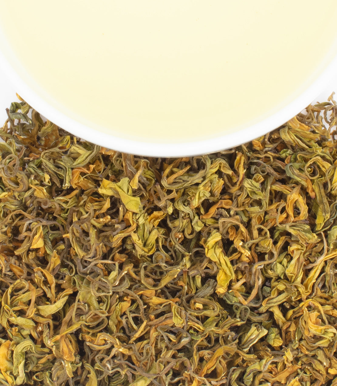 A cup of pale yellow tea sits above a pile of dried, curly green and yellow tea leaves.