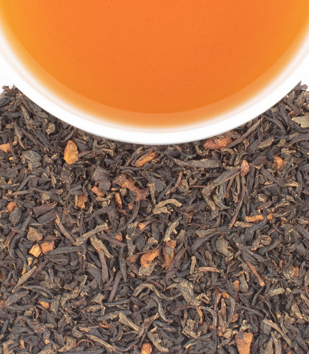 Close-up of loose-leaf tea with dark brown to black leaves and small orange-brown pieces, with a white cup rim and amber tea visible at the top.