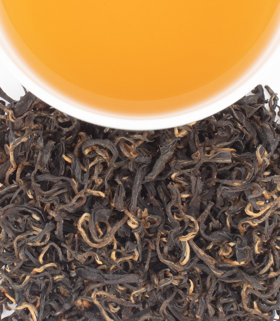 A cup of golden tea sits above a pile of dark, curly loose-leaf tea against a white background.