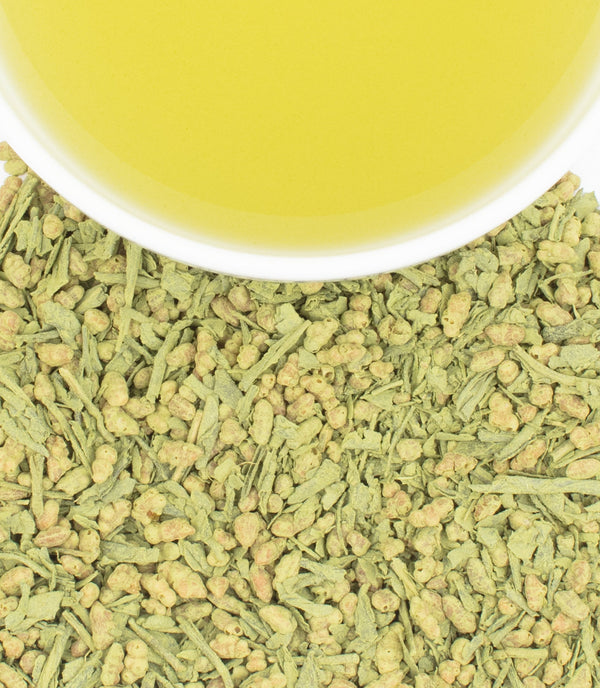 A cup of pale yellow tea rests atop a bed of loose green tea leaves and rice coated in matcha