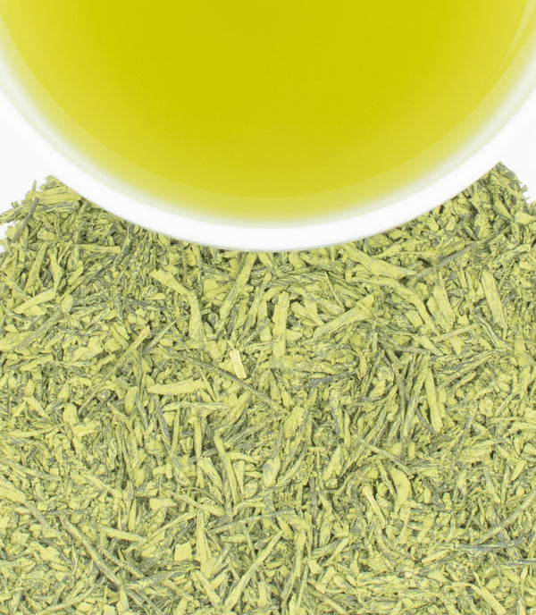 A cup of bright green tea sits above a pile of dried green tea leaves against a white background.