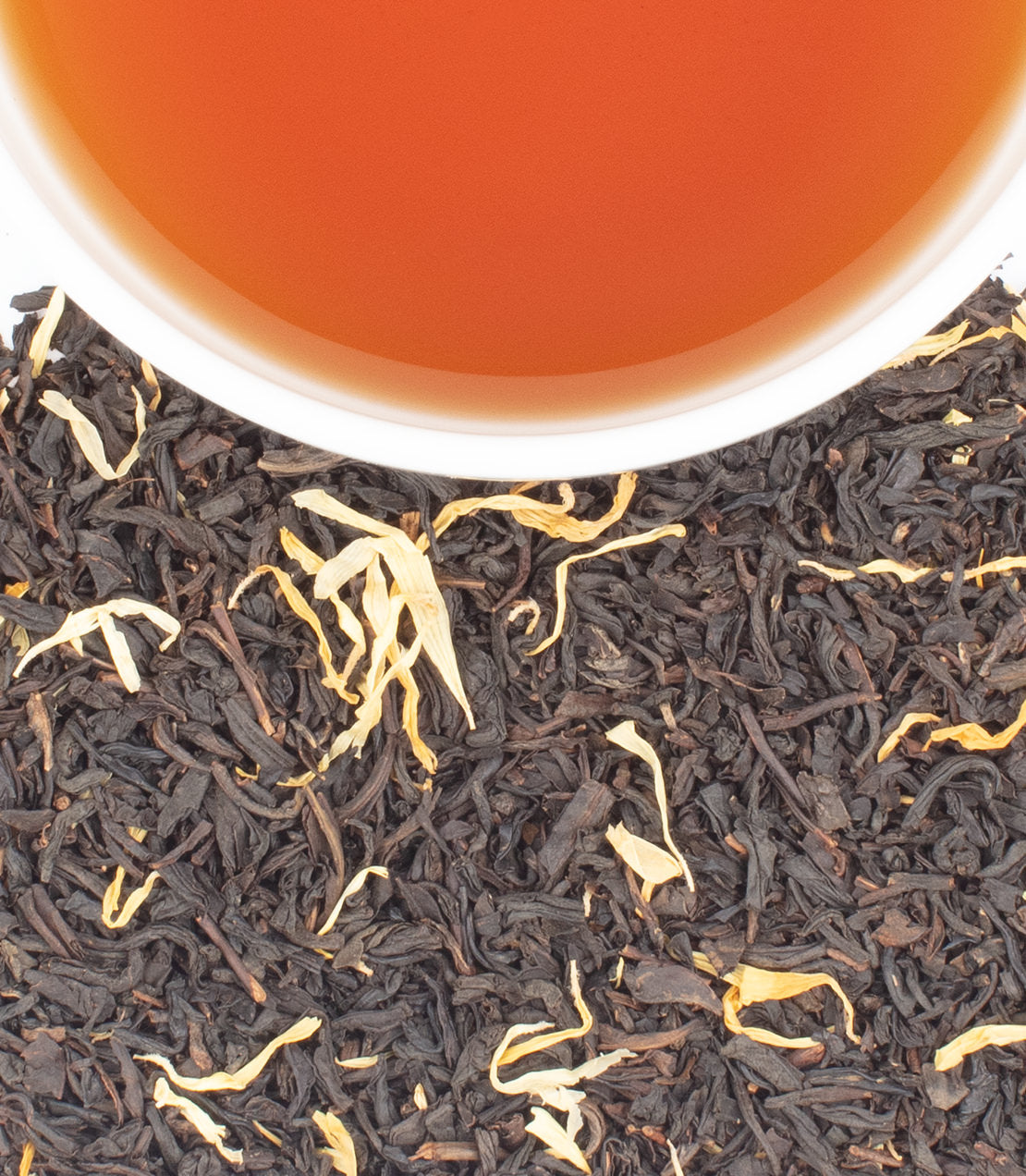 Close-up of loose-leaf tea with dark brown to black leaves and a few pale yellow floral tips, with the rim of a white cup filled with amber tea at the top.