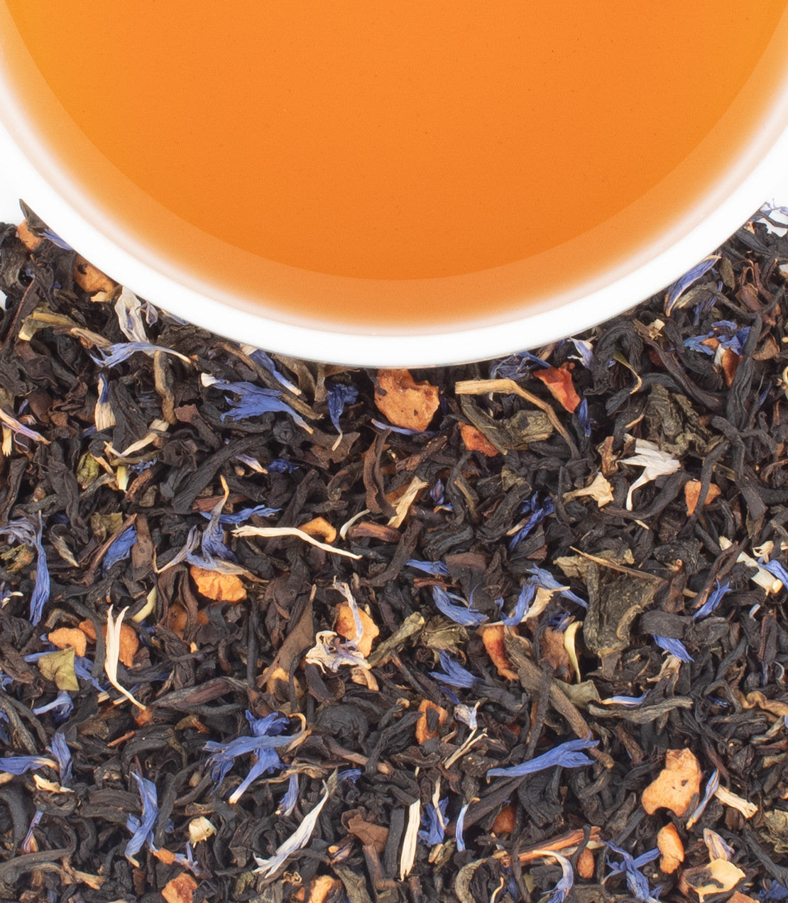 A cup of amber-colored tea sits above a spread of dark, colorful loose-leaf tea with blue and orange floral pieces.