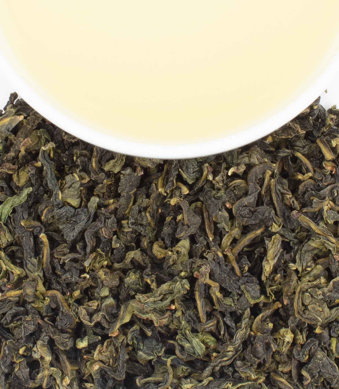 A white cup filled with pale tea sits above a pile of dark, twisted tea leaves.