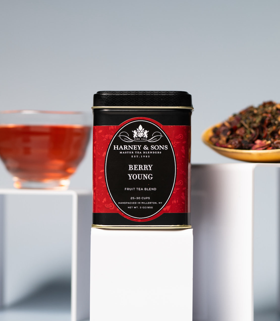 Harney & Sons Berry Young tea tin with a glass of tea and a bowl of tea leaves on a gray background.