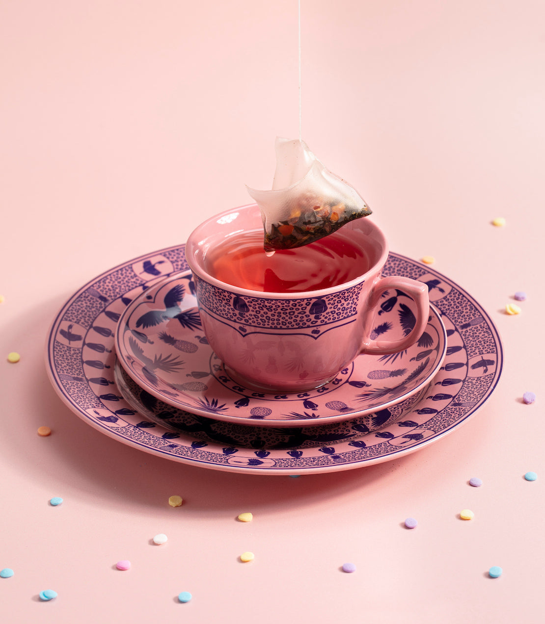 Pink teacup with a tea bag on a decorative plate against a pink background with colorful polka dots.
