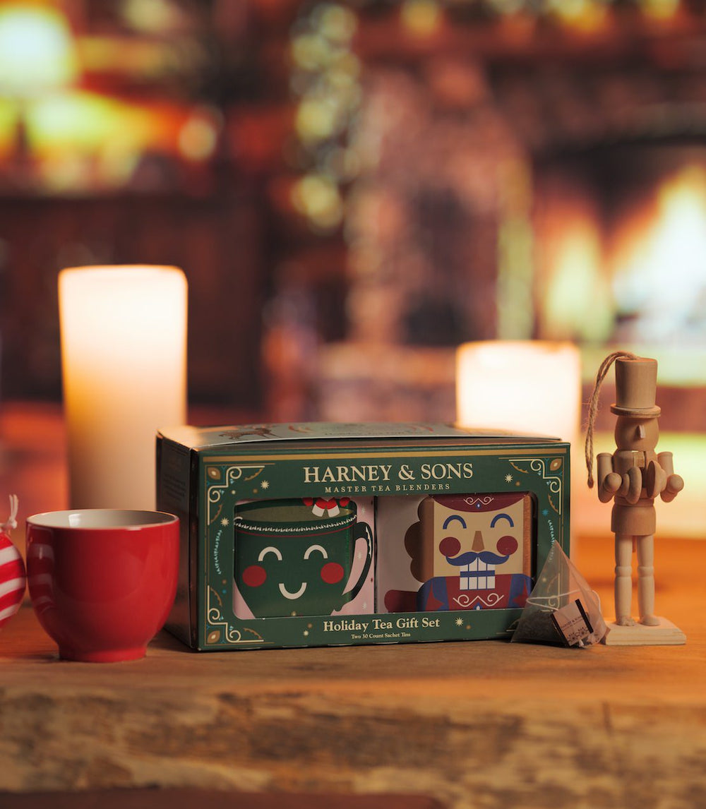 Harney & Sons Holiday Tea Gift Set in a green box, styled on a wooden table with a red mug, a nutcracker, and a red-and-white striped candy cane against a warm, blurred holiday background. Gift box includes Nutcracker and Candy Cane teas, in 30 count tins.