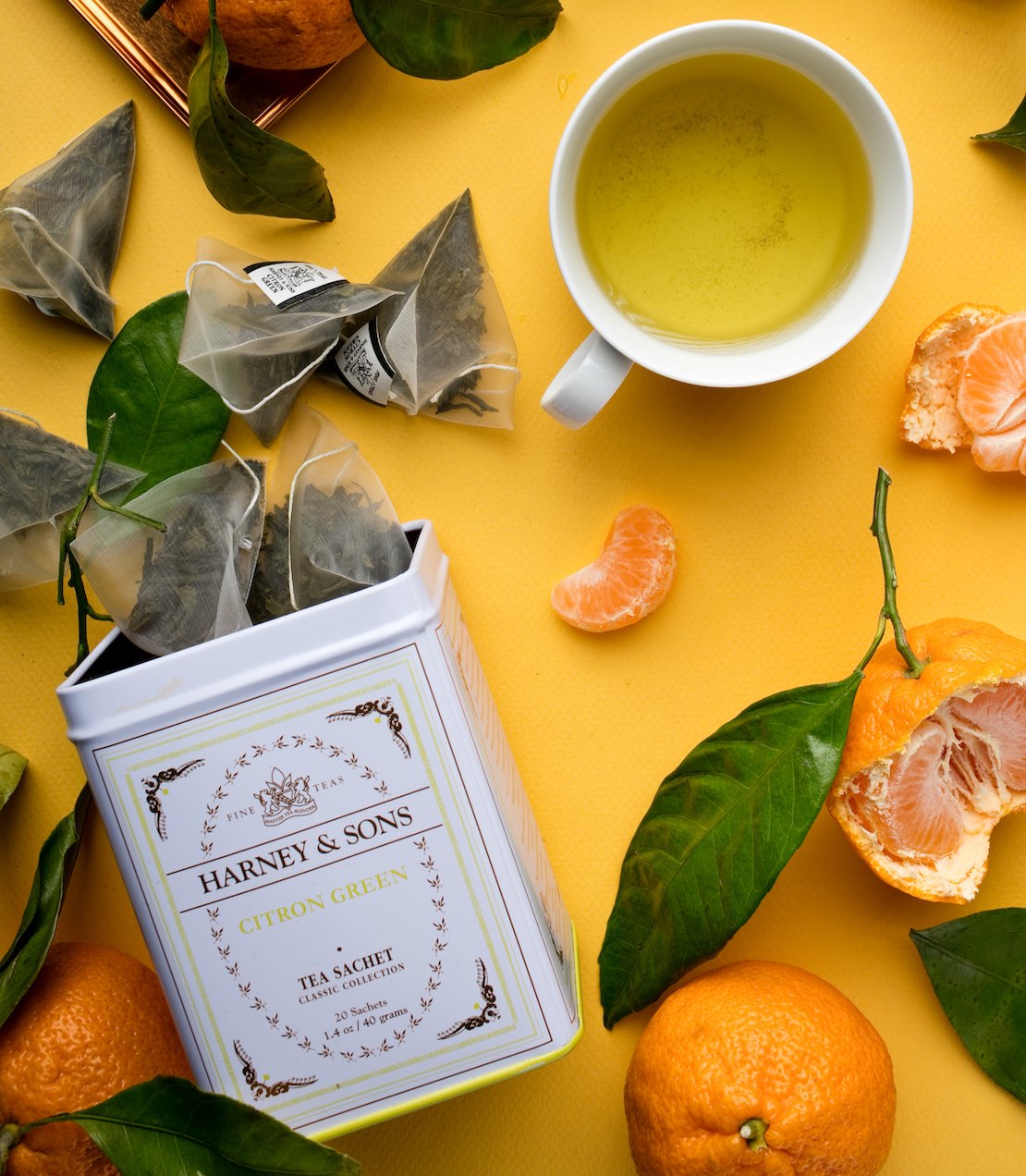 Tin of Citron Green Classic Sachets, a cup of tea, and oranges on a yellow background.