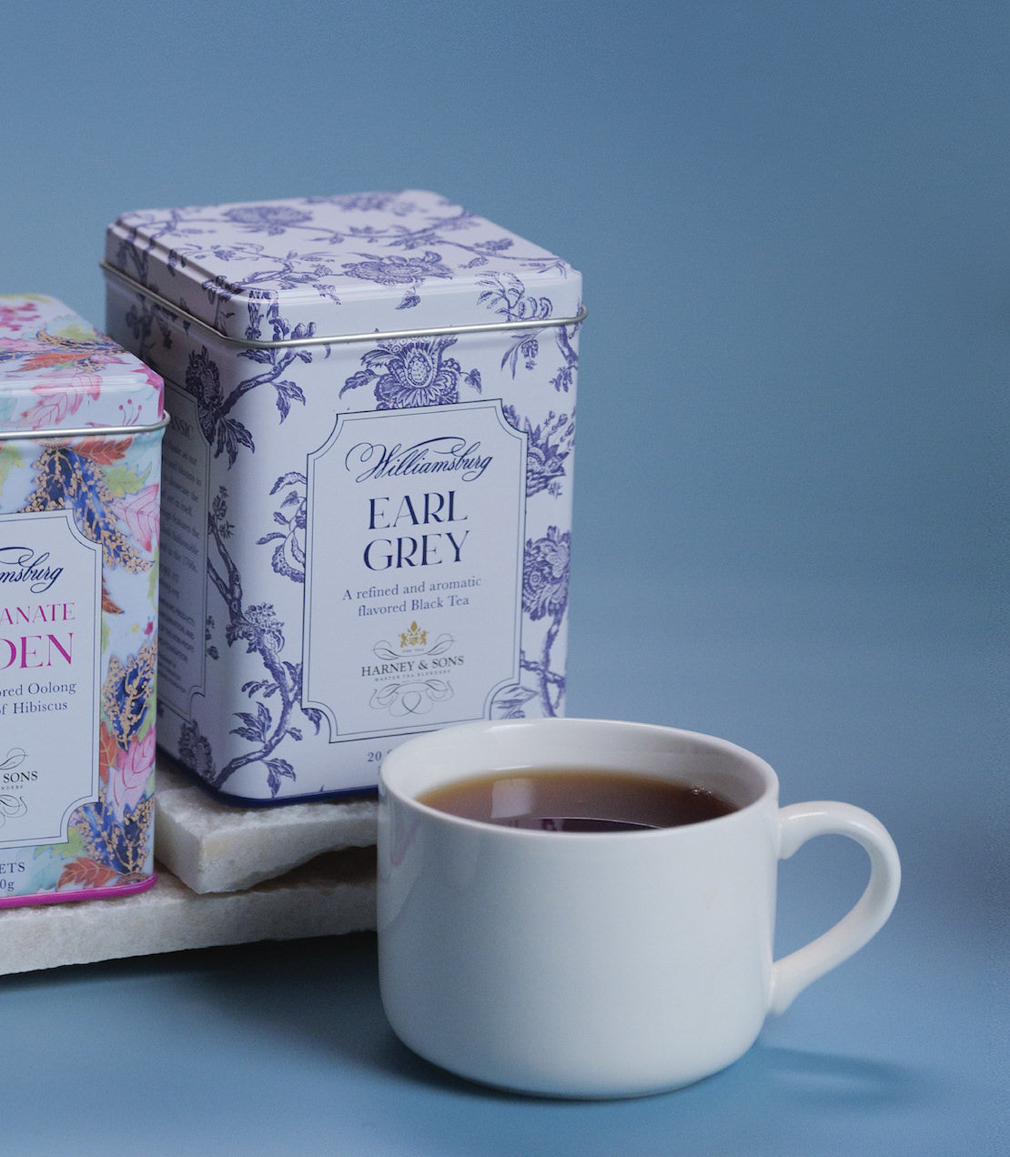 Williamsburg Earl Grey tea tin with 20 sachets beside a cup of brewed tea on a light blue background.