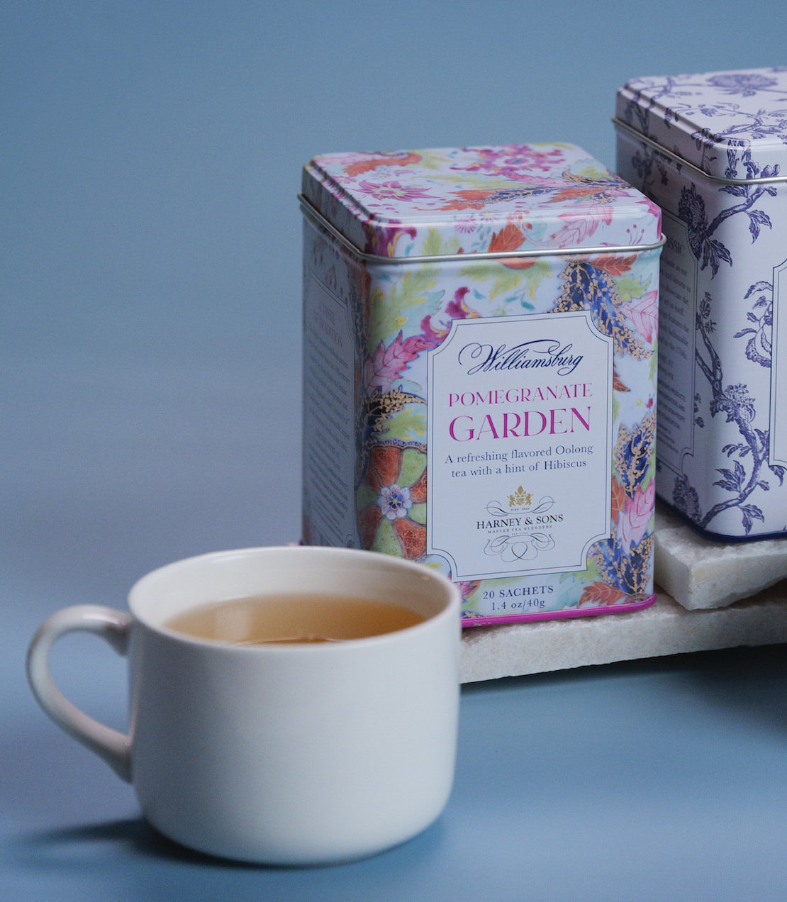 Williamsburg Pomegranate Garden tea tin with 20 sachets beside a cup of brewed tea on a light blue background.