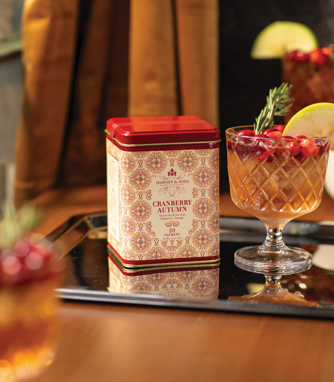 Cranberry Autumn tea tin with 20 sachets beside a glass of cranberry drink garnished with lime and rosemary.