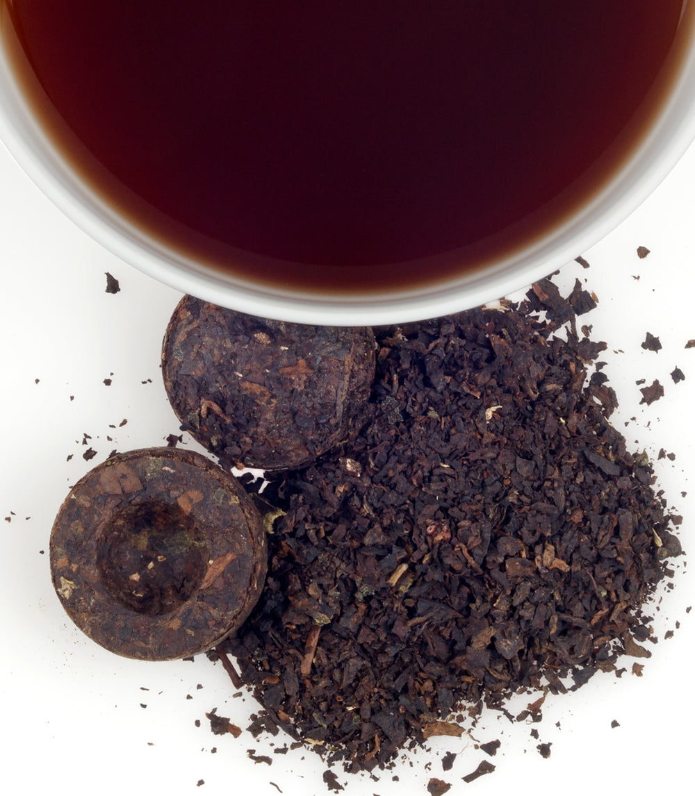 2015 Nuo Mi Pu-Erh tea with loose leaves and compressed tea cakes beside a cup of dark brewed tea.