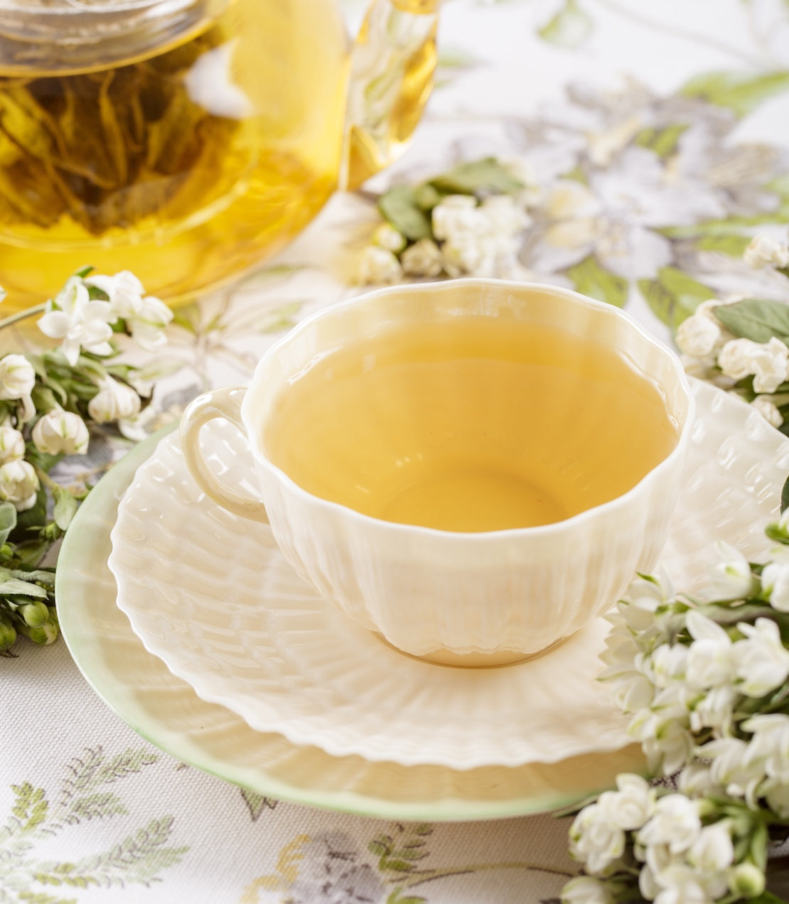 White teacup filled with yellow tea on a matching saucer, surrounded by white flowers on a light background.