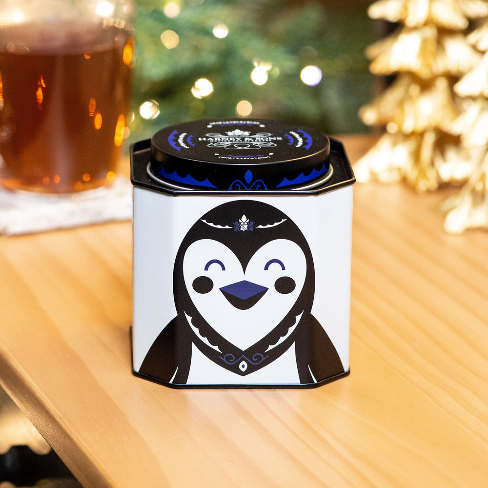 Tea canister with penguin design on a wooden surface with blurred lights in the background
