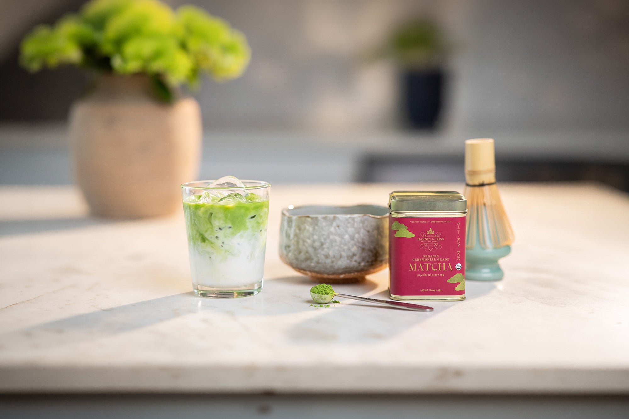 A styled product shot of a matcha tea set on a light marble countertop. The set includes a glass of prepared matcha iced tea, a pink and gold 'The Matcha' tin, a textured gray bowl, a bamboo whisk, and a small scoop, with a blurred background showing a vase of green leaves.