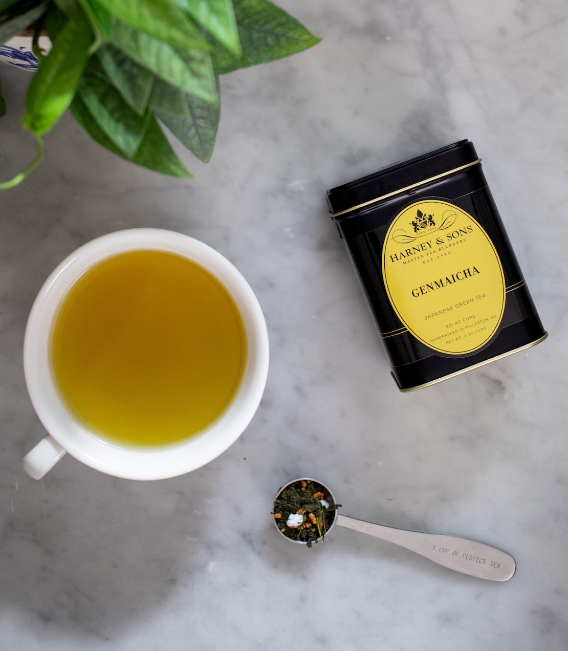 Tea cup with brewed tea, a tea tin of Harney & Sons Genmaicha tea, and a spoonful of tea leaves on a marble surface.