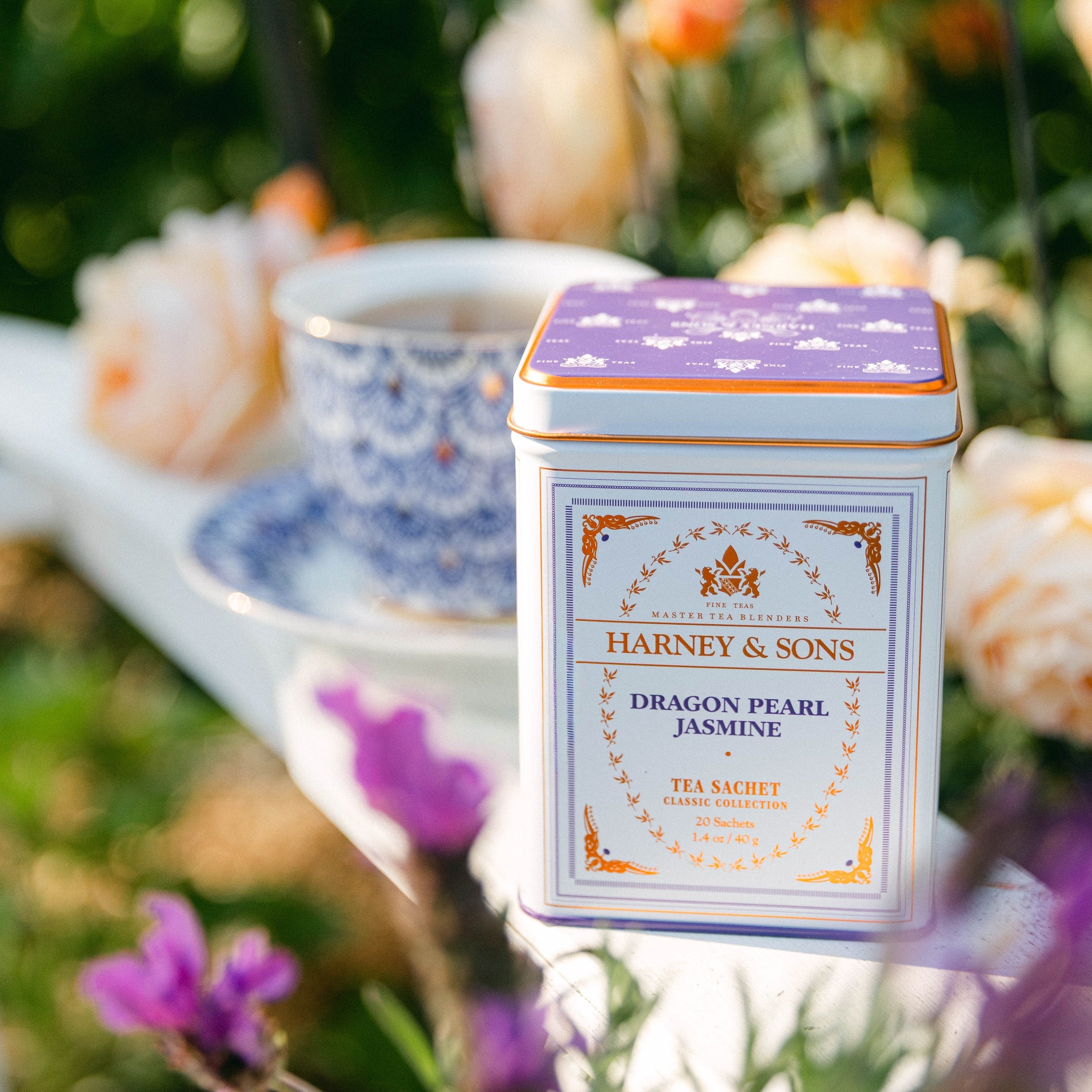A square metal tea tin from Harney & Sons, Dragon Pearl Jasmine, set in a beautifully blurred garden setting with a teacup in the background.