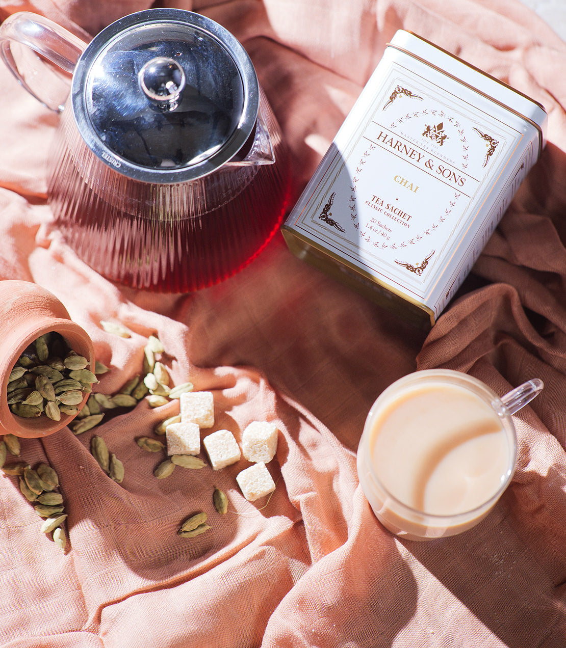 Classic tin of Harney & Sons chai sachets with a teapot, glass of chai, and spices on a textured fabric background.
