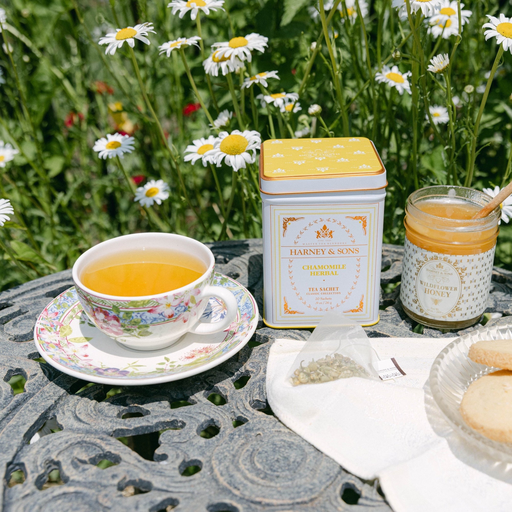 A tea and cookie spread arranged on a round metal garden table outdoors, with a cup of tea, a tin of honey, a jar of honey with a wooden dipper, and cookies on a plate, all set against a background of green grass and white daisies.