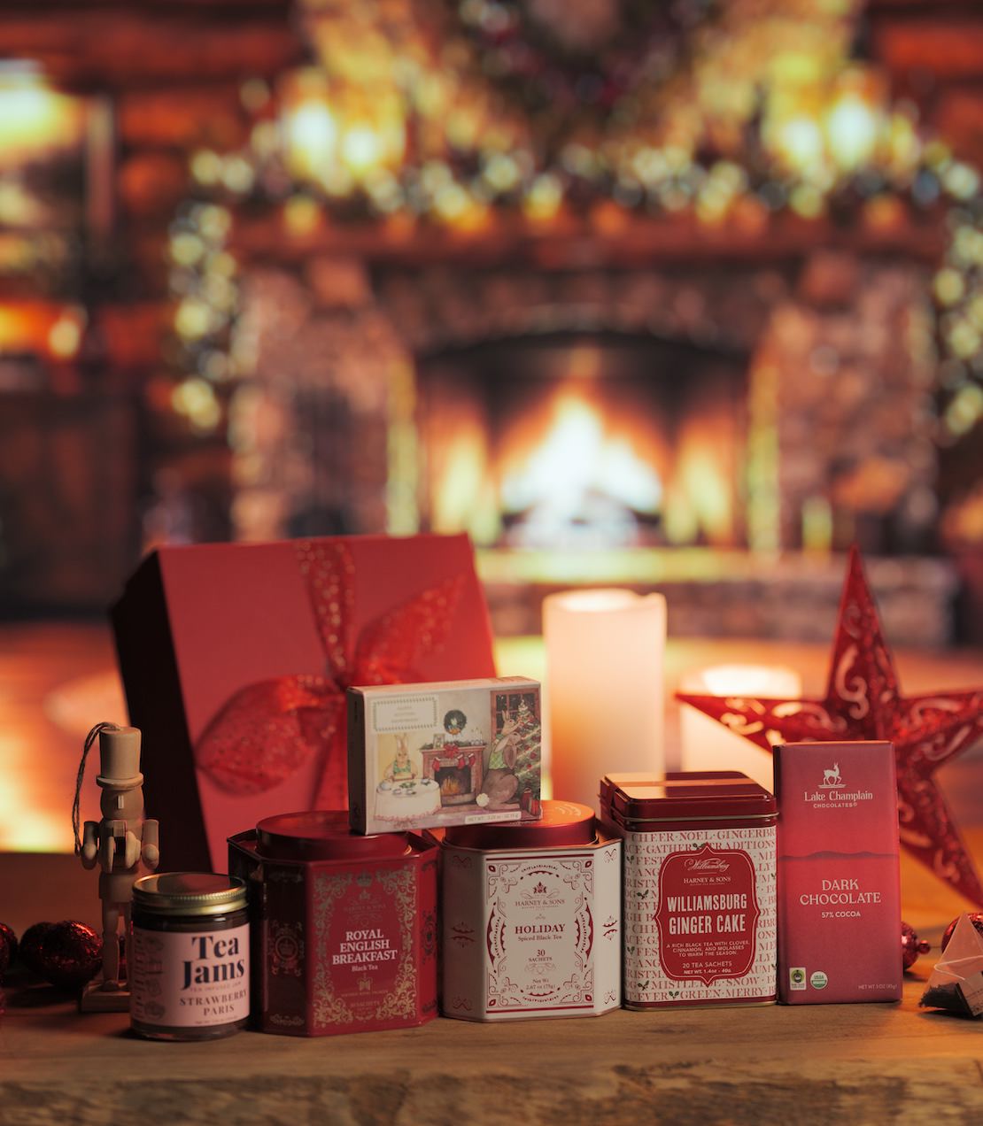 A curated holiday tea and chocolate gift set arranged on a wooden surface in front of a warmly lit fireplace. The set includes a red gift box, tea tins, shortbread, a tea jam.