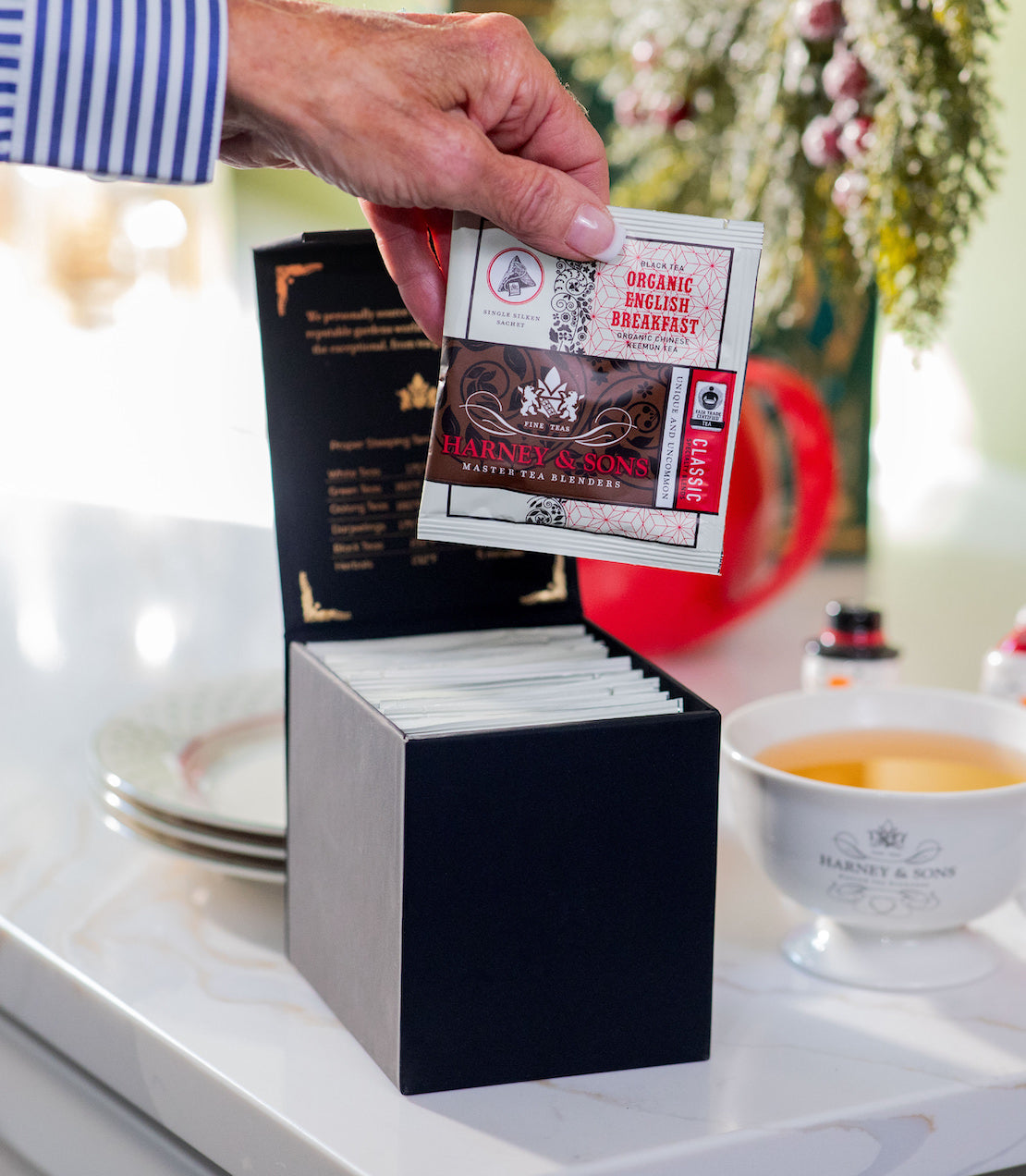Person holding a tea bag over an open black box of tea bags on a kitchen counter.