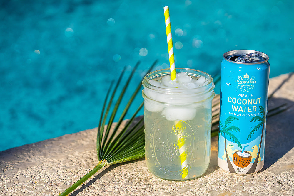 Harney & Sons Premium Coconut Water served over ice in a mason jar with a paper straw beside the can, poolside.