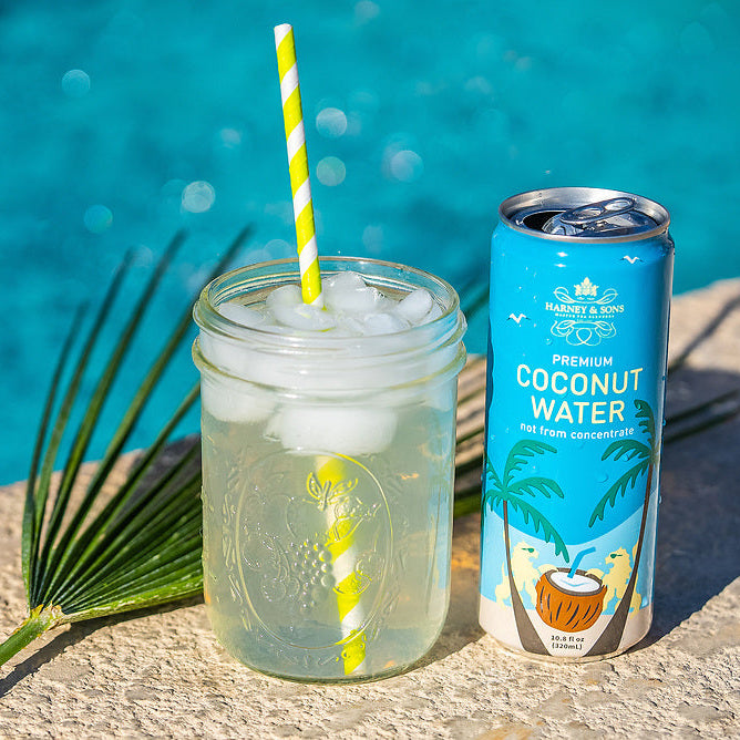 Harney & Sons Premium Coconut Water served over ice in a mason jar with a paper straw beside the can, poolside.