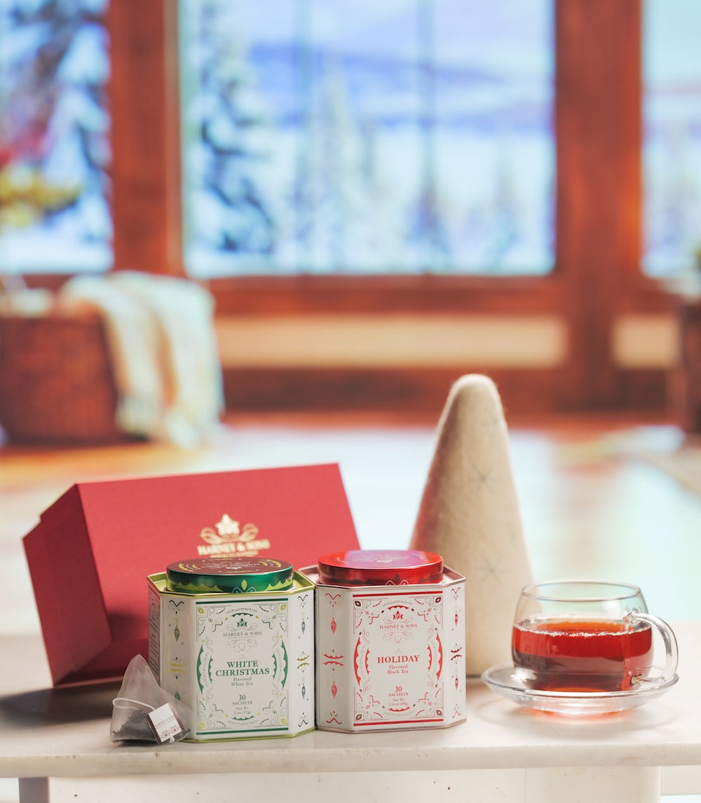 A red and blue Royal Holiday tin sits on a marble surface in front of a warm, blurred fireplace. A glass teacup filled with tea and a tea bag are on a saucer nearby, with plums and greenery in the foreground.