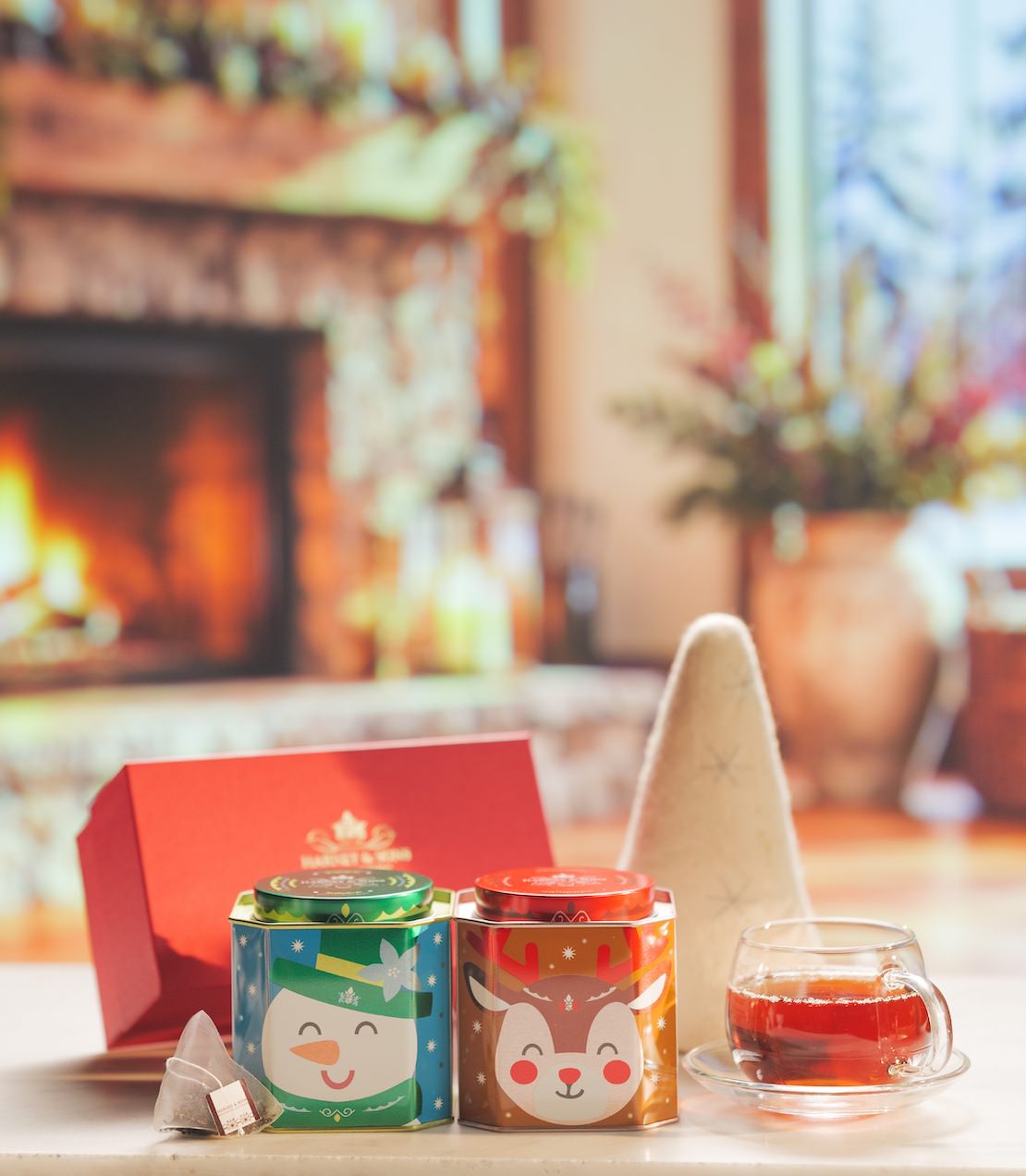 Two Christmas-themed metal tea tin cans with festive illustrations of a snowman and a reindeer, shown with a red gift box, tea sachets, and a glass cup of tea on a table in front of a warm, blurred fireplace background.