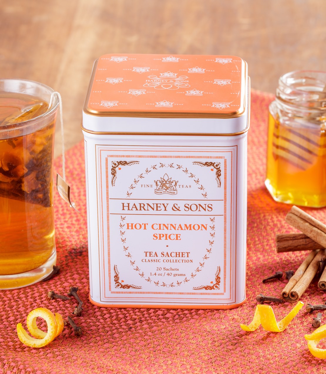 Harney & Sons Hot Cinnamon Spice tea tin with a glass of tea and cinnamon sticks on a red surface.