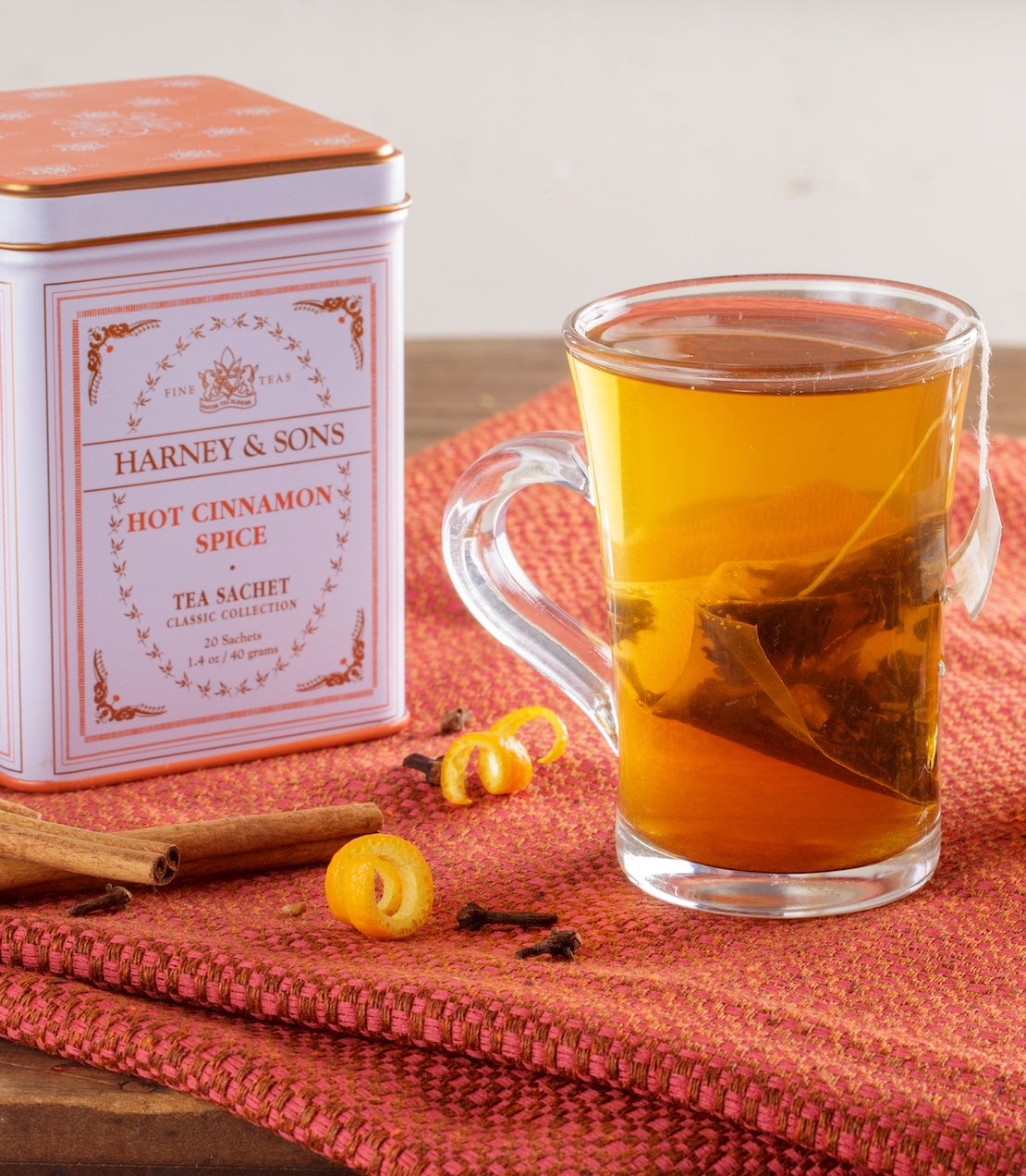 Harney & Sons Hot Cinnamon Spice tea tin with a glass of tea and cinnamon sticks on a red surface.