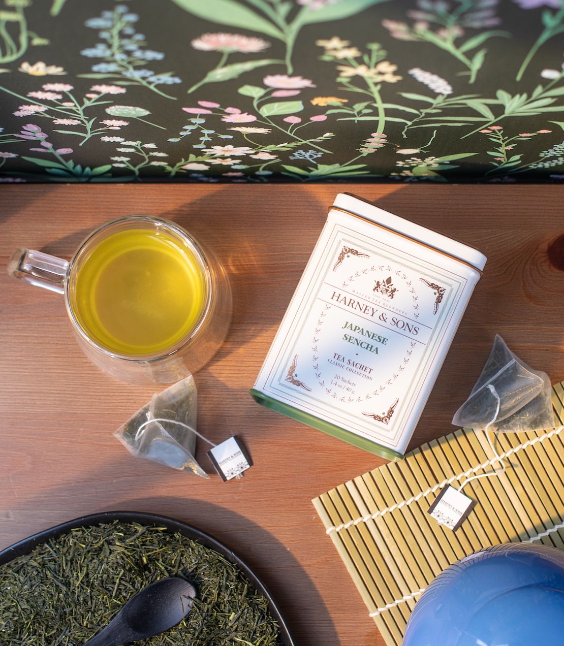 Tea-making setup with Harney & Sons Japanese Sencha Classics tea tin, a cup of green tea, and tea leaves on a wooden surface.