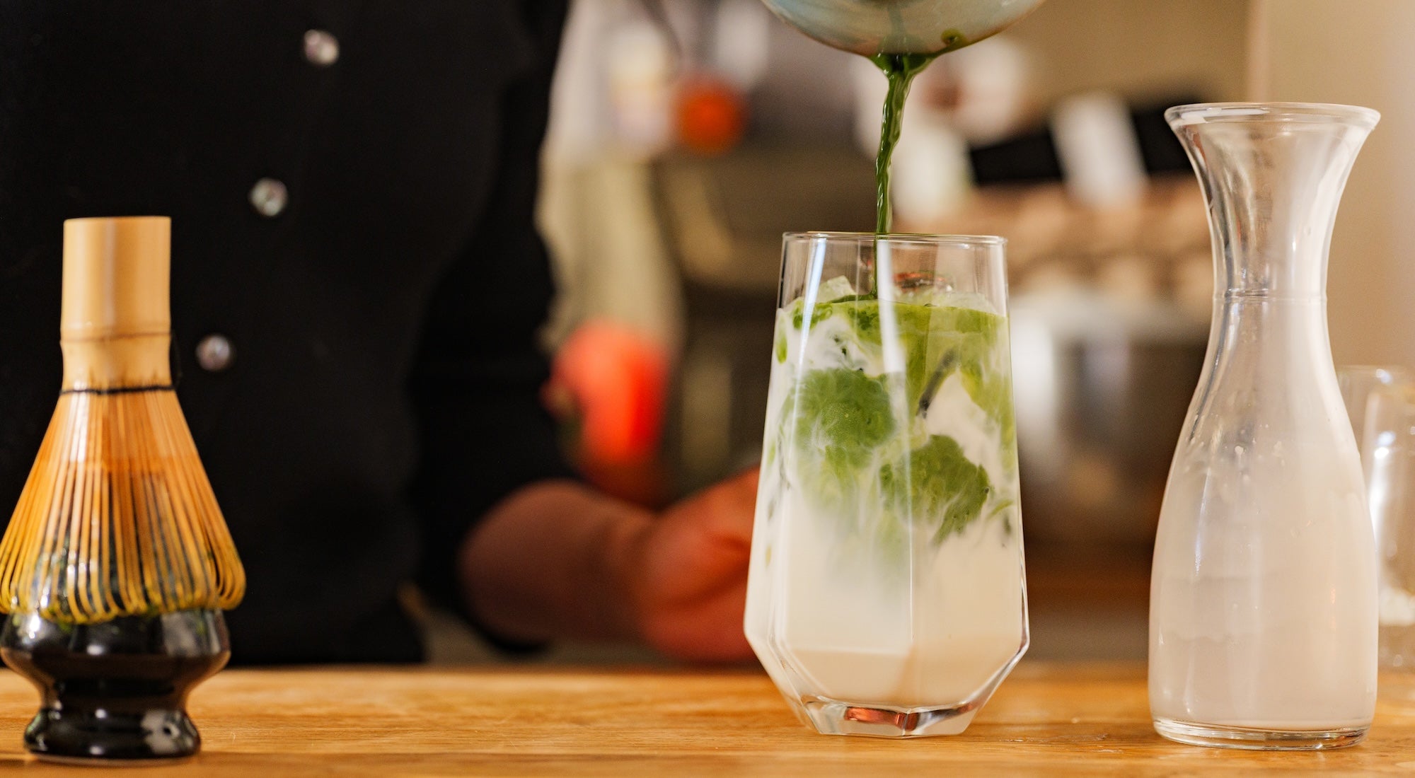 A person in a black uniform pours green tea into a clear glass filled with white milk or foam, set on a wooden table with a bamboo whisk and a glass carafe nearby.