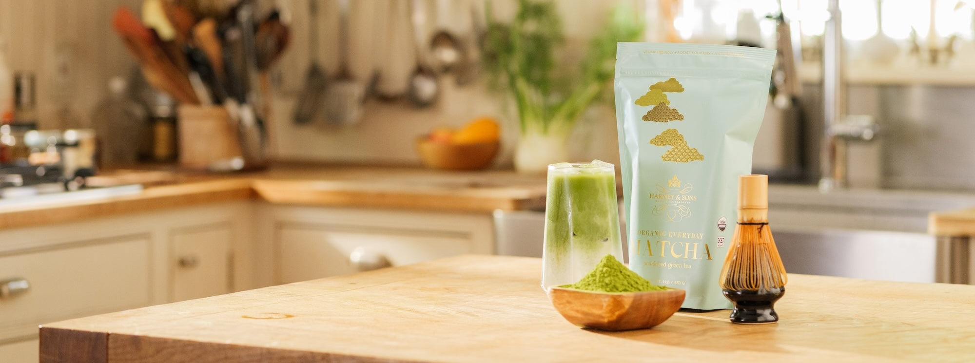 A pouch of Avichia matcha green tea powder, a glass of brewed green tea, a small wooden bowl filled with matcha powder, and a bamboo whisk are arranged on a wooden countertop in a softly blurred kitchen setting.