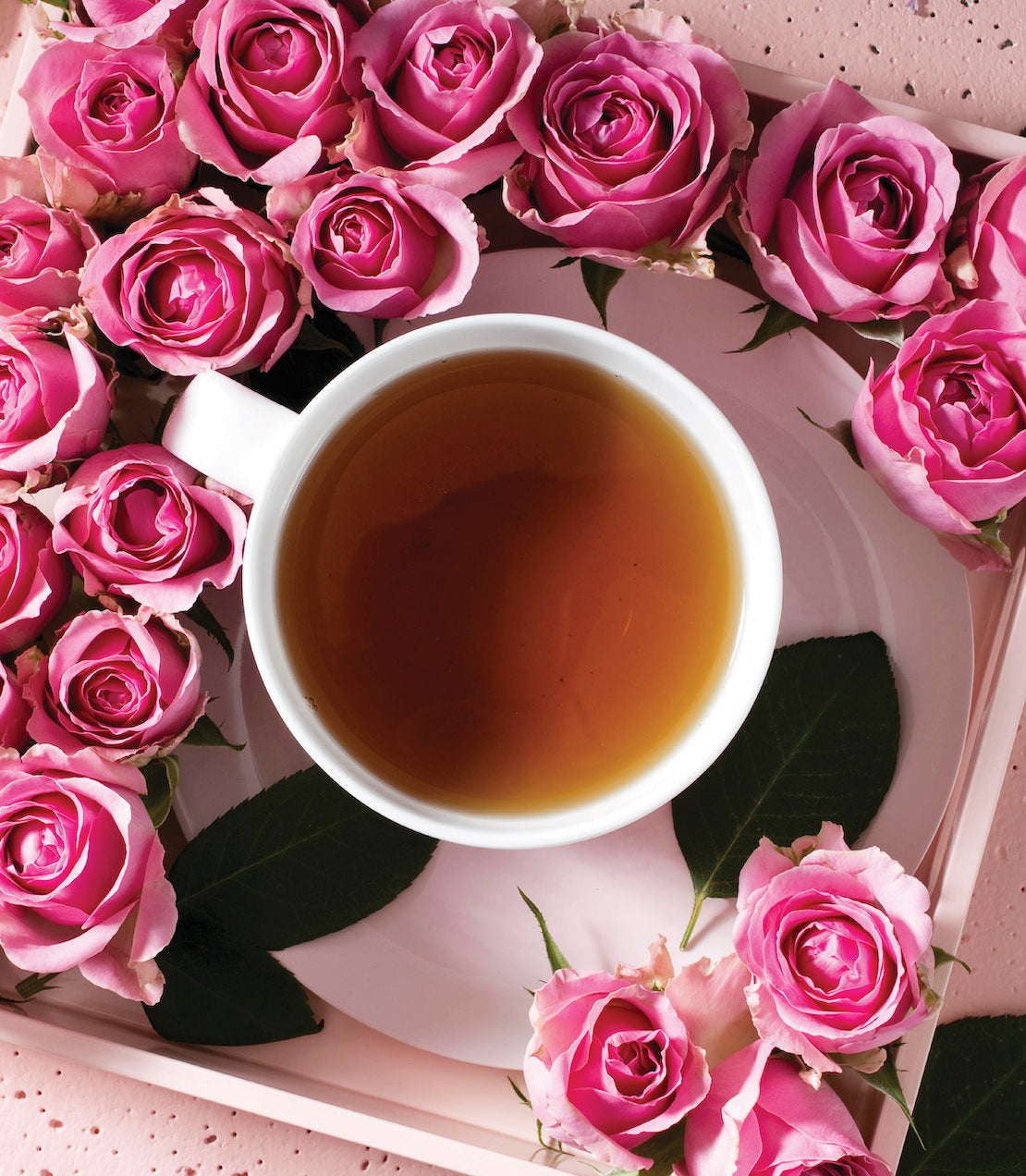 A white cup of rose-scented tea surrounded by fresh pink roses on a pastel background.