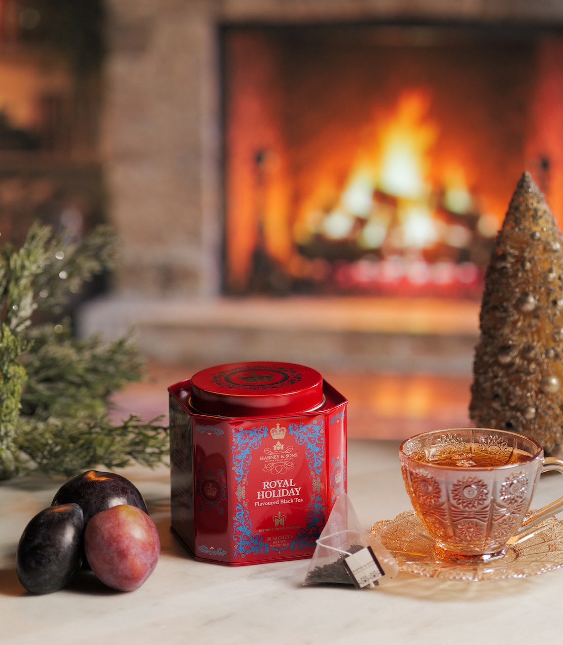 A red and blue Royal Holiday tin sits on a marble surface in front of a warm, blurred fireplace. A glass teacup filled with tea and a tea bag are on a saucer nearby, with plums and greenery in the foreground.