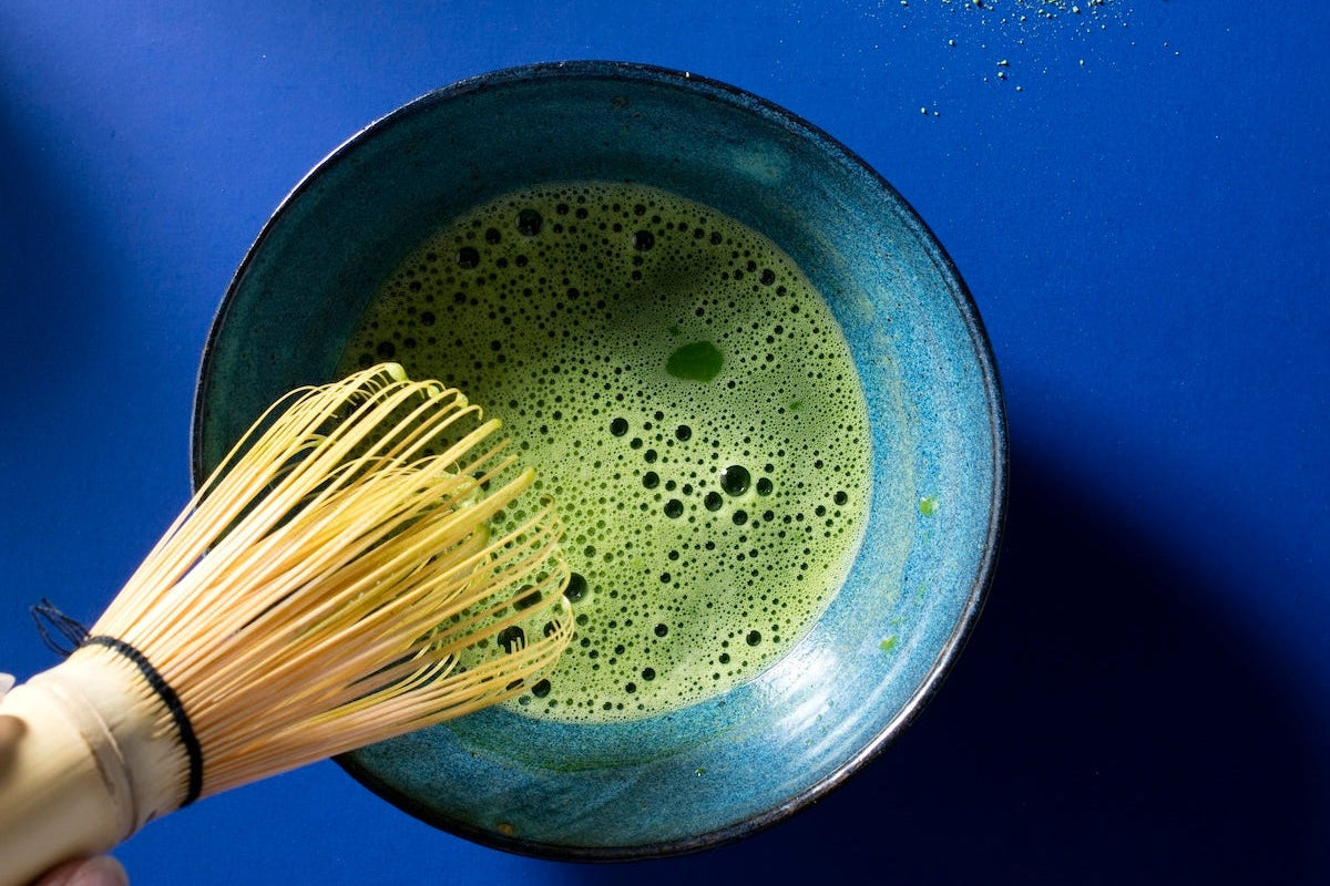Matcha green tea being whisked in a bowl with matcha powder on a blue background