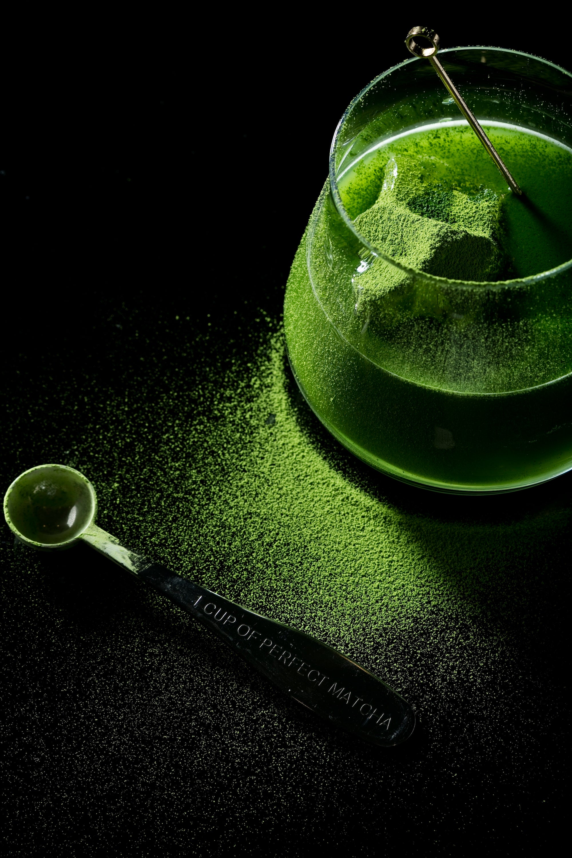 A glass bowl filled with bright green matcha powder and a metal spoon resting inside, with a scoop of matcha powder placed beside it on a dark surface. The spoon is engraved with '1 CUP OF PREMIUM MATCHA'.