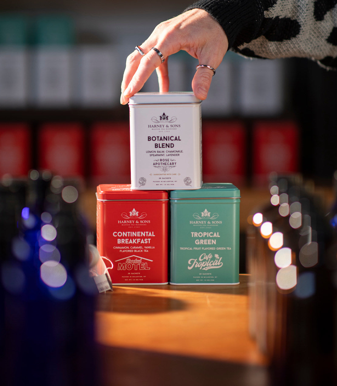 Hand holding a tin of Botanical Blend tea with Continental Breakfast and Tropical Green tea tins on a blurred background.