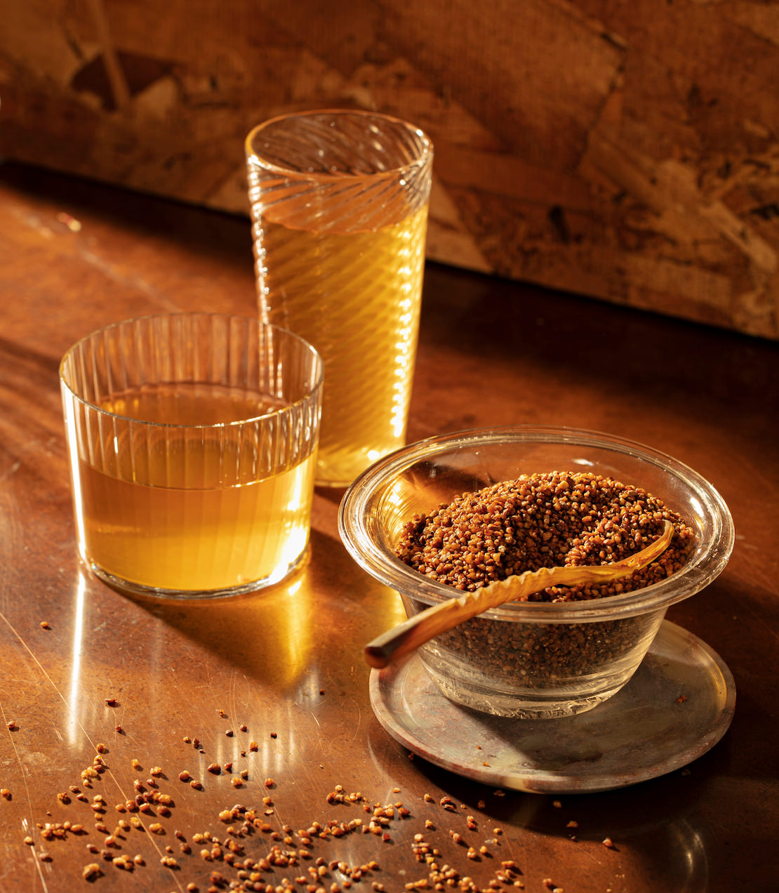 Soba roasted buckwheat in a glass bowl with a wooden spoon, alongside two glasses of golden liquid on a wooden table.