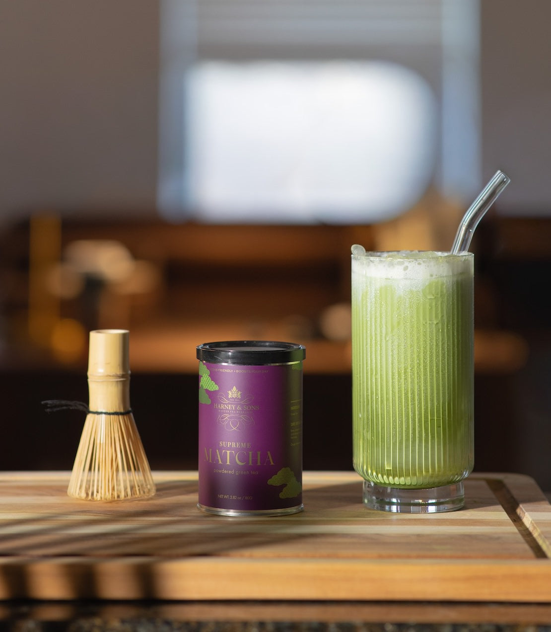Matcha green tea tin, bamboo whisk, and glass of matcha drink on a wooden surface.