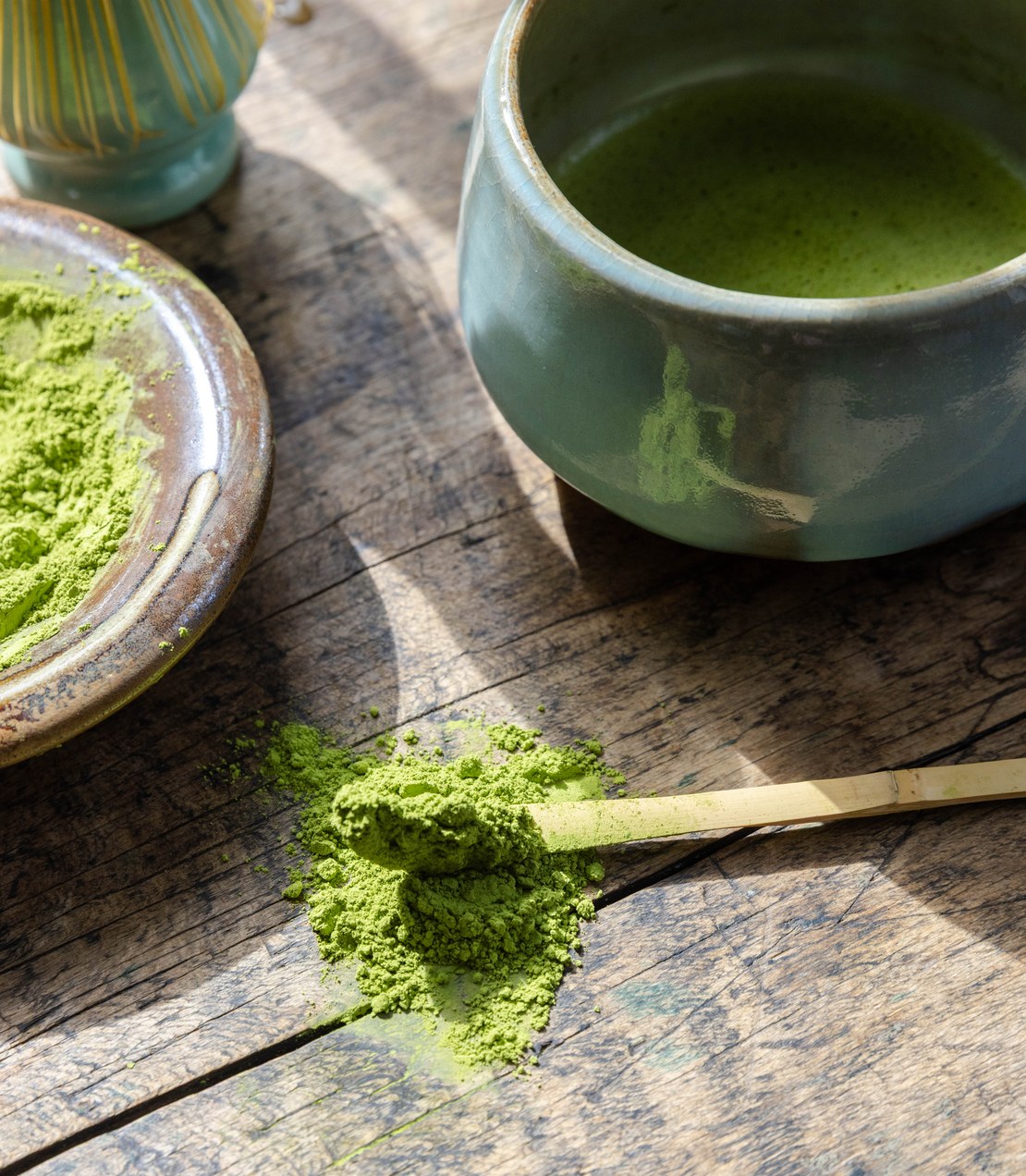 A bowl of whisked Supreme Matcha with a dish of matcha powder and a bamboo scoop on a weathered wooden table.