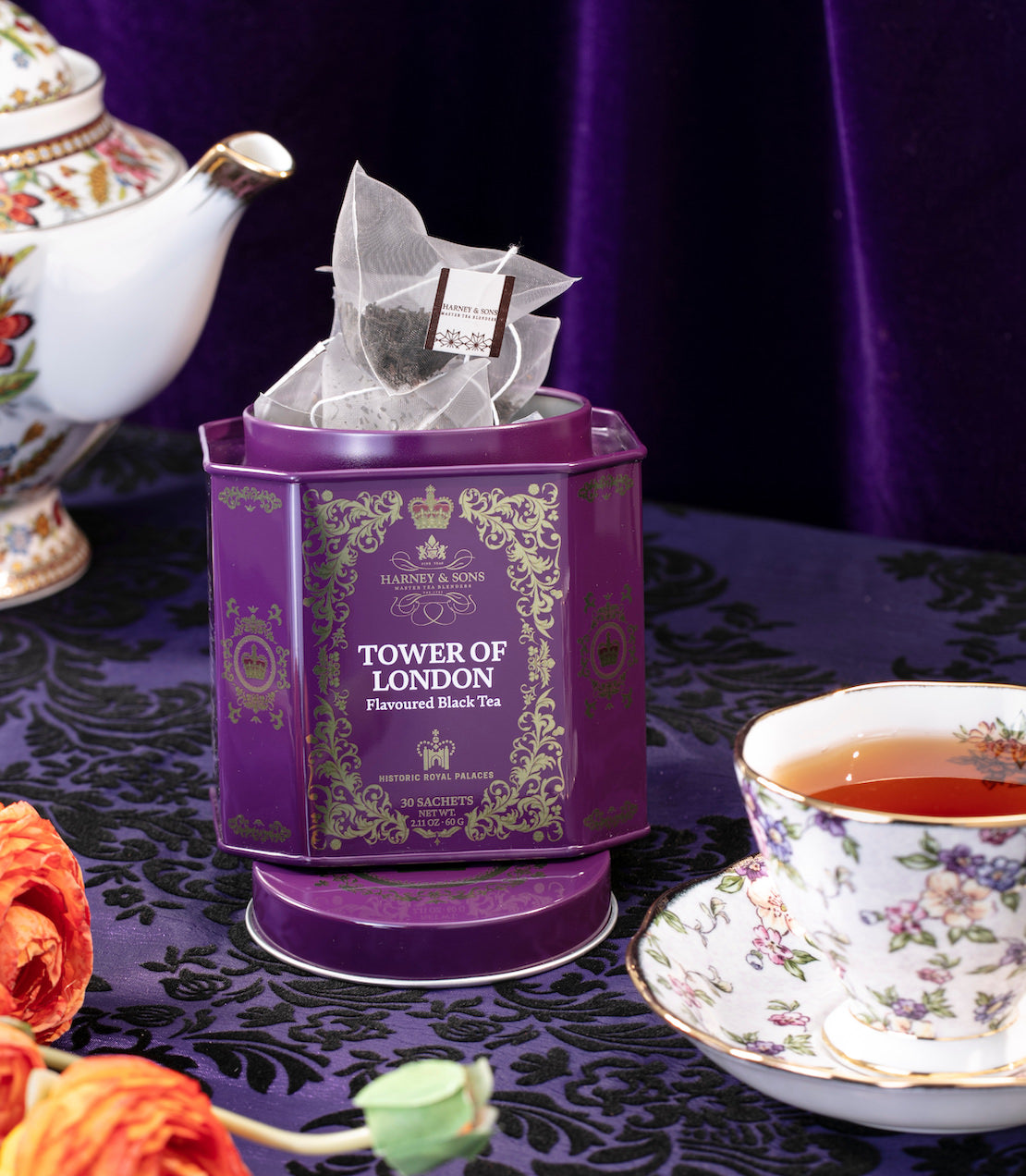 Tower of London tea tin on a table with a floral and purple tablecloth, teapot, and teacup. 