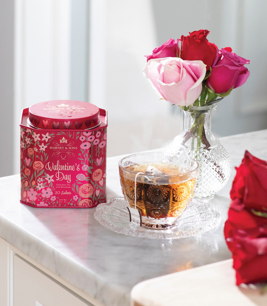 Valentine's Day tea tin with 30 sachets beside a glass cup of tea and a vase of pink and red roses.