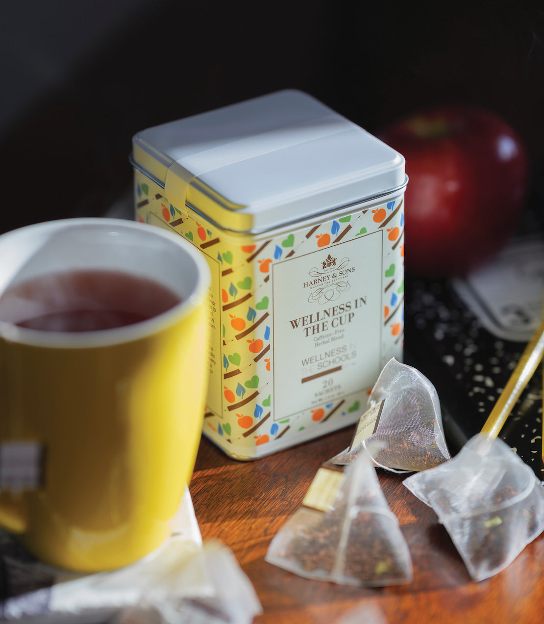 Tin of 20 Wellness in the Cup sachets beside a yellow mug of tea and an apple on a wooden table.