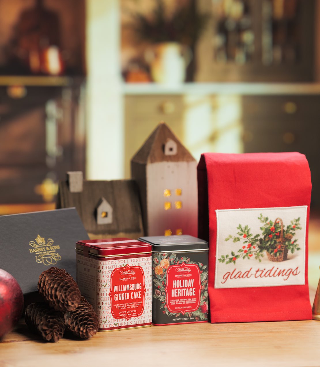 A holiday gift set on a wooden surface featuring a red tea towel labeled 'glad tidings', two decorative metal tins, a dark gift box, pinecones, and a blurred warm-toned indoor background.