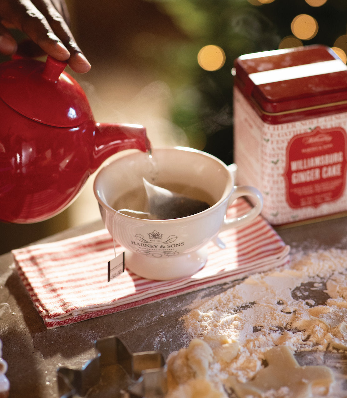 Pouring hot water from a red teapot into a cup with a sachet of Williamsburg Ginger Cake tea, with festive decorations in the background.