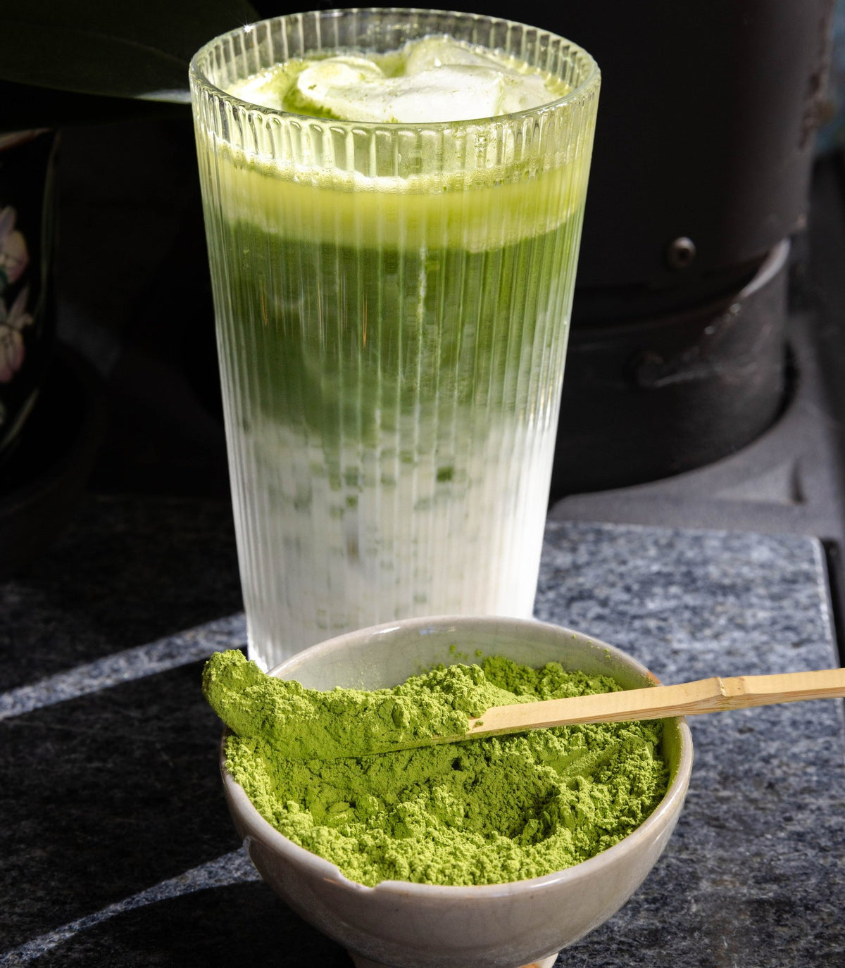 A layered iced matcha latte in a ribbed glass with a bowl of matcha powder and a bamboo scoop.